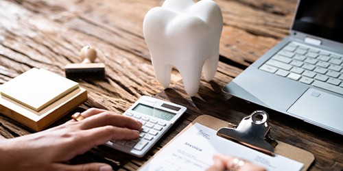 Hand on calculator and holding pen to write on invoice at desk with model tooth, stamp pad, and laptop