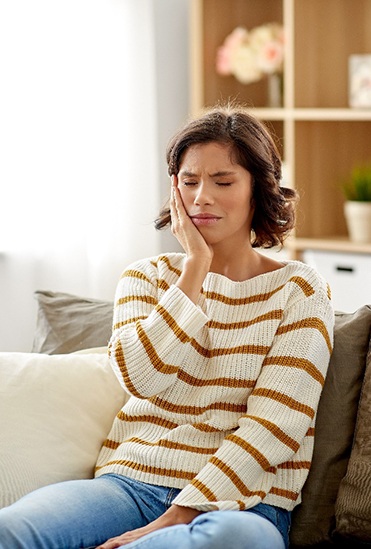 Woman sitting on couch and cupping her cheek due to toothache